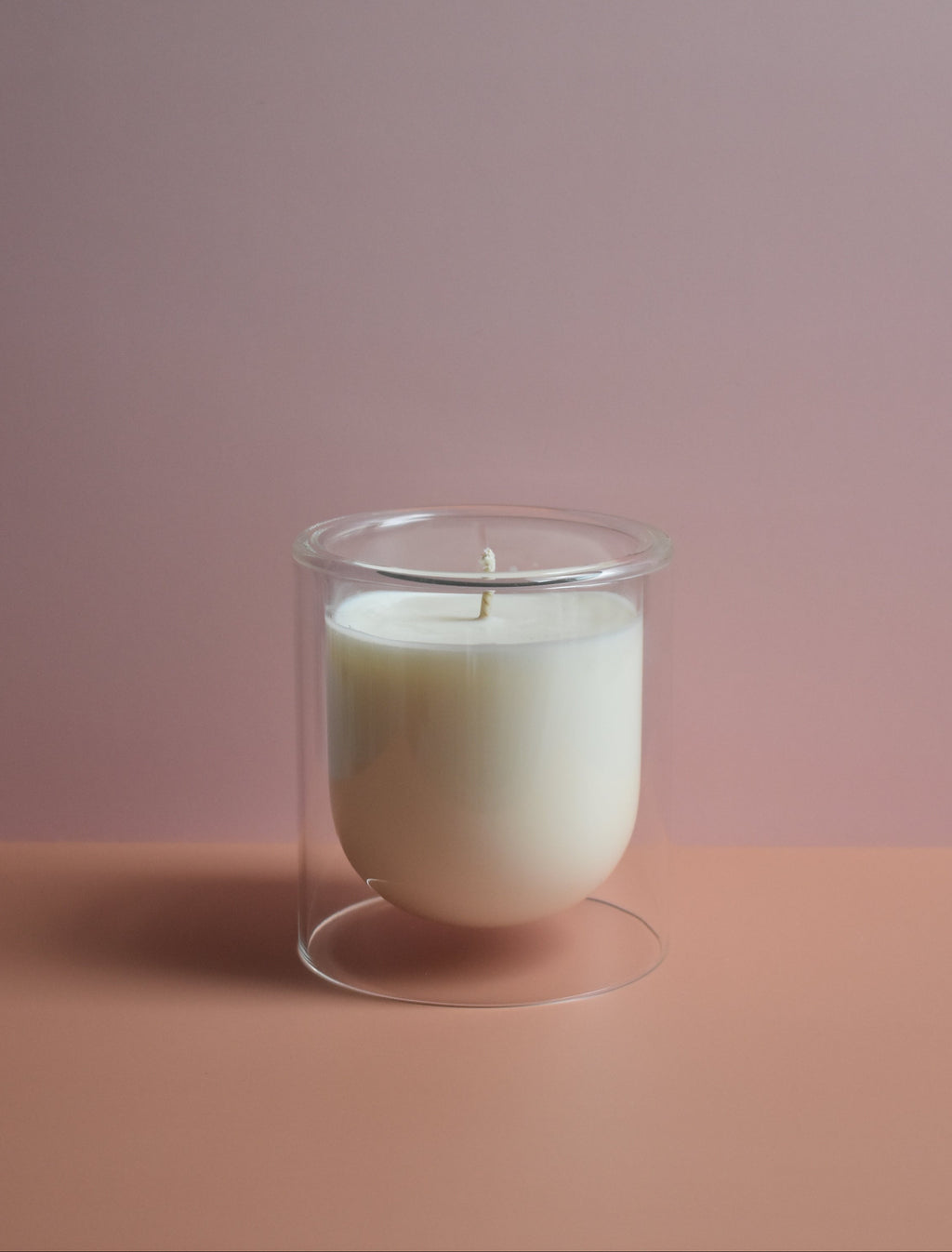 White candle in a clear glass holder on a pink surface with a plain background